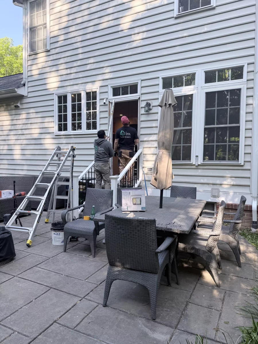 Two workers installing a door on a house exterior with ladder and patio furniture visible.
