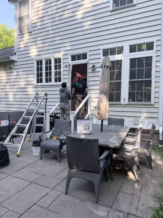 Two workers installing a door on a house exterior with ladder and patio furniture visible.