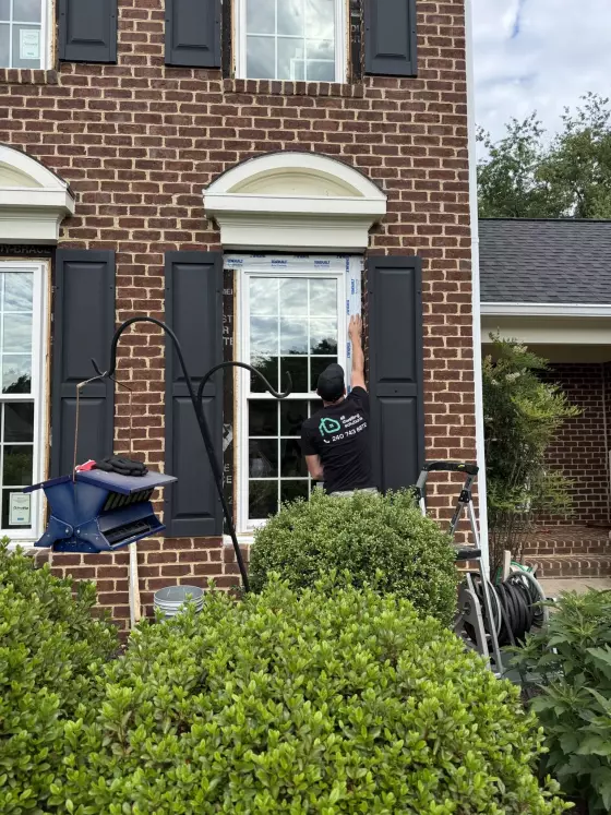 Worker installing window on brick house exterior with shutters and ladder
